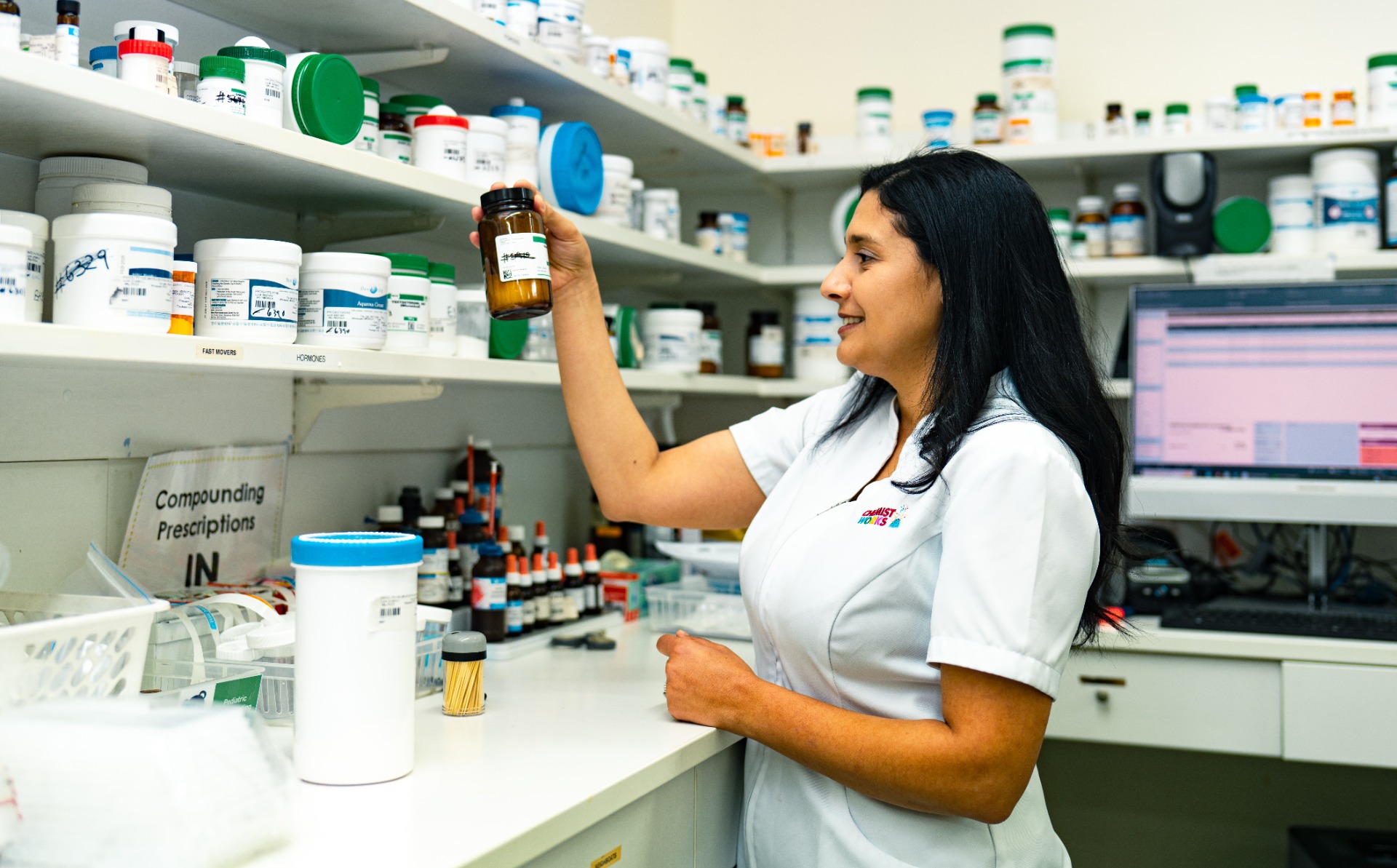 Pharmacist organising medication in a compounding pharmacy Female pharmacist in a white coat smiling while organising prescription bottles in a compounding pharmacy with shelves of medication and a computer workstation in the background.